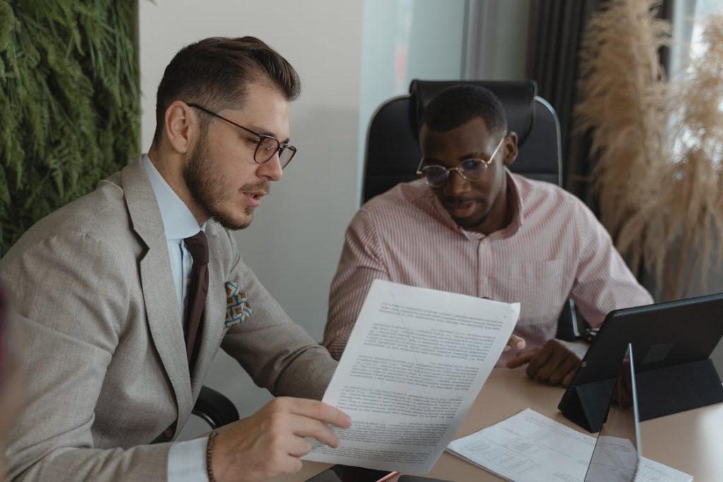 Two businessmen reviewing a document together with laptops on a table.